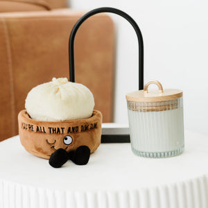 A plush bun-shaped decorative object with embroidered text and black pom-pom feet sits on a white table beside a ribbed glass jar candle with a wooden lid and black metal stand in a softly lit indoor setting.