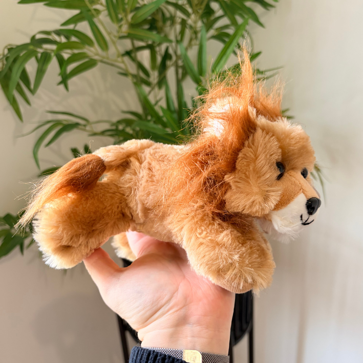 A small plush lion toy with fluffy tan and cream fur is being held in a person's hand in front of a white wall and green plant leaves.