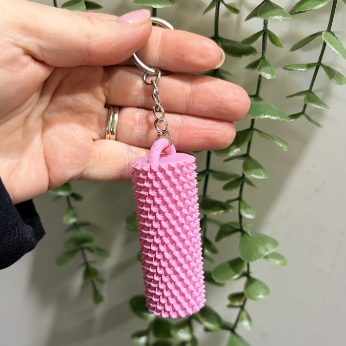 A pink textured cylindrical keychain held by a hand, featuring a spiky, pleated design and a silver metal keyring, with a green trailing plant in the background.