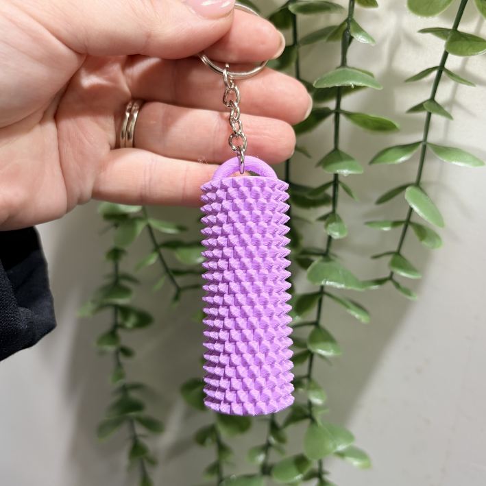 A hand holding a lavender purple textured cylindrical keychain with a silver split ring, set against a background of green trailing houseplants.