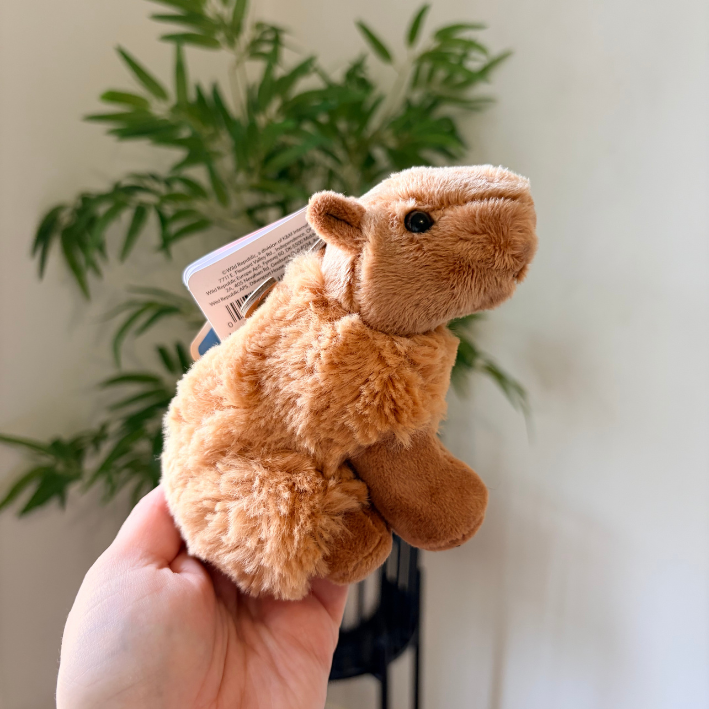 A small plush camel toy being held in a person's hand, with a paper tag attached and a blurred indoor plant in the background.