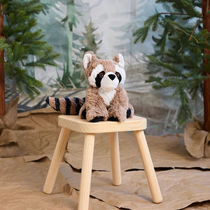 A plush raccoon toy sits on a small wooden stool in a staged forest scene with artificial trees and a blurred woodland backdrop.