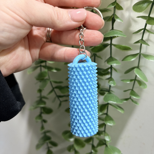 A light blue textured cylindrical keychain held by a hand, featuring a silver metal key ring and chain, with a green trailing plant in the background.