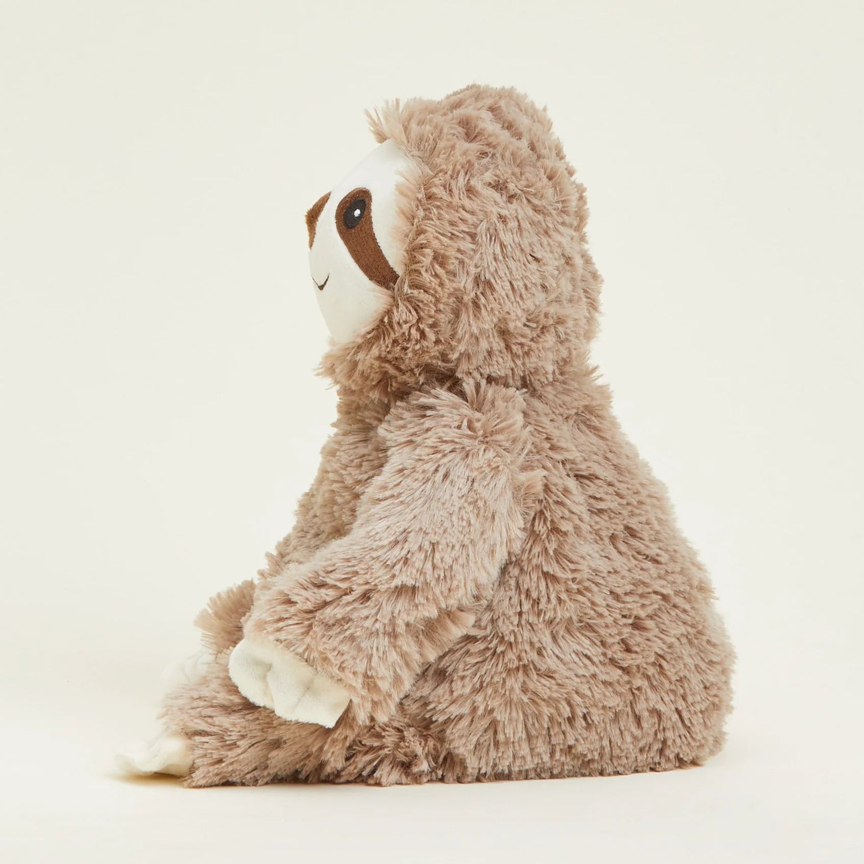 A plush sloth toy sitting in profile, featuring shaggy light brown fur, a white face, and dark brown facial details, photographed against a clean off-white background.